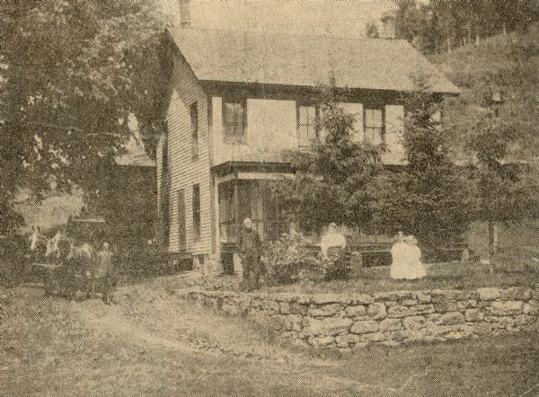 Exterior view of the Salisbury house, with a horse-drawn wagon located to the left side of the home and William Salisbury, John Salisbury, Mrs. William Salisbury and their daughters Velma and Beulah standing from left to right in front of the house. A steep hill is in back of the house on the right.