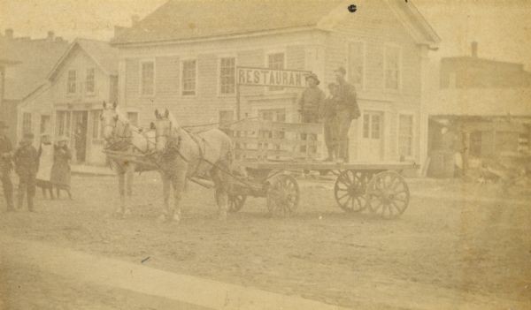 View of a street scene featuring people standing on a horse-drawn wagon in the foreground, and a restaurant in the background.