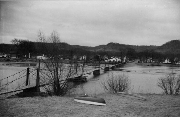 View of a footbridge over the Pine River. Boats are laying in the grass in the foreground.