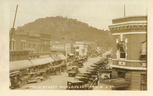 Elevated view of Main Street with automobiles lining either side.  An ice cream parlor is visible on the right side, as are a dry cleaning and tailoring business, a tire shop, and a large Coca-Cola billboard along the left. Caption reads: "Main Street, Richland Center, Wis."