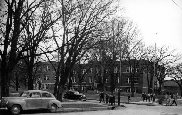 Exterior view towards a high school, with students walking on the sidewalks and several parked automobiles.