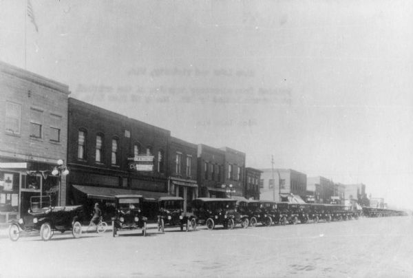View across street towards a long row of automobiles parked on the left side at an angle along the curb in front of storefronts.