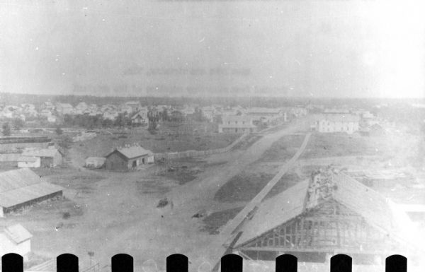 Elevated view of Rice Lake. A partially constructed building is in the right foreground. Unpaved roads and a sidewalk lead to commercial buildings and dwellings in the distance.