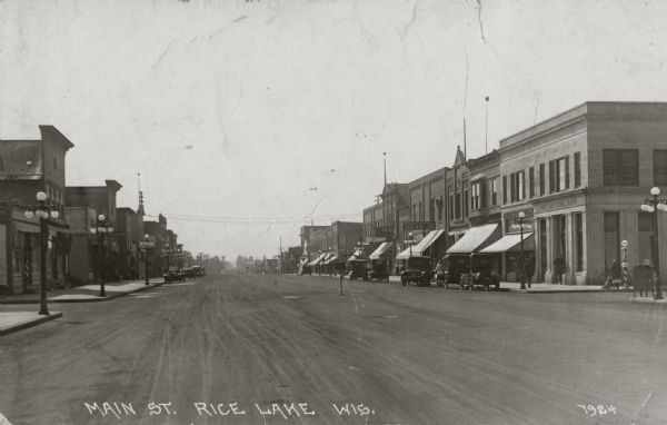 View down Main Street. Storefronts are along the sidewalks on both sides of the street, and lampposts and cars are along the curb.