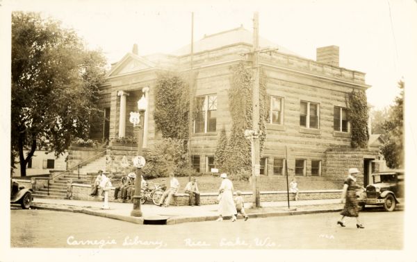 View of Carnegie Library on the corner of South Main Street and West Messenger, Rice Lake. Pedestrians are crossing the street, automobiles are parked along the curb, and people are sitting on the stone wall along the sidewalk.