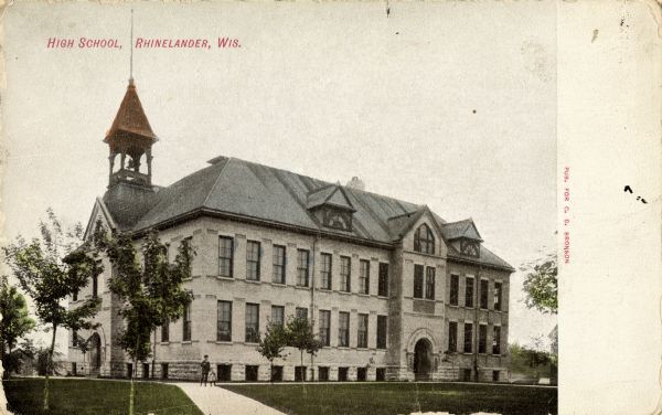 View up sidewalk toward the high school. A man and a child are standing on the sidewalk in front. Caption reads: "High School, Rhinelander, Wis."