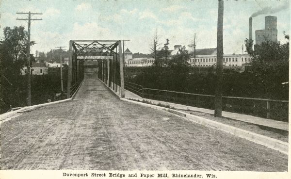 View down center of the Davenport Street Bridge with a paper mill in the distance on the right. Caption reads: "Davenport Street Bridge and Paper Mill, Rhinelander, Wis."