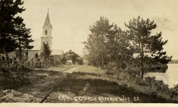 View towards a Catholic church further down a fence-lined road on the left. On the right is a shoreline. Caption reads: "Catholic Church Reserve Wis."