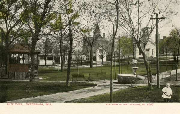 View of the city park, with a fountain and benches, and a child standing in the foreground on the right. Caption reads: "City Park, Reedsburg, Wis."