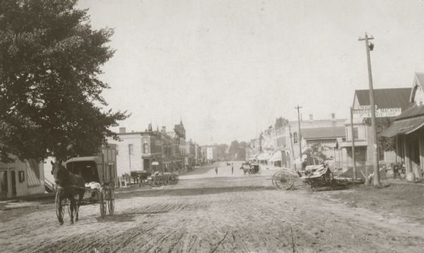 View down unpaved Main Street, with a machine and repair shop on the right and multiple horse-drawn vehicles traveling along the road.
