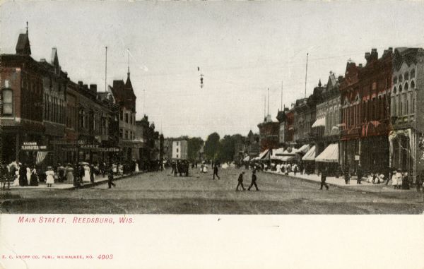 View looking down Main Street. Pedestrians are on the sidewalks, and walking across the street. Caption reads: "Main Street, Reedsburg, Wis."