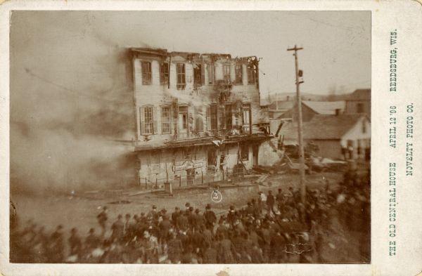 Elevated view of a burning Central House engulfed in smoke, with a crowd of spectators gathered to watch. Text on side reads: "The Old Central House April 12 '96 Reedsburg, Wis."