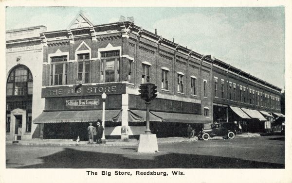 Exterior view from intersection towards The Big Store on the corner, with two men standing outside the entrance. A large traffic signal is in the center of the street. Caption reads: "The Big Store, Reedsburg, Wis."