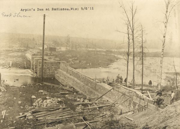 Elevated view of construction on Arpin's dam. Men are working on the right. Caption reads: "Arpin's Dam at Radisson, Wis".
