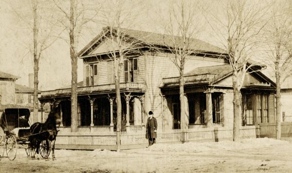 The Utly Residence, surrounded by a fence, on the north-east corner of Wisconsin and 8th Streets. Built by E.R. Cooley. A man is standing in the center near the corner, and a horse-drawn buggy is on the far left.