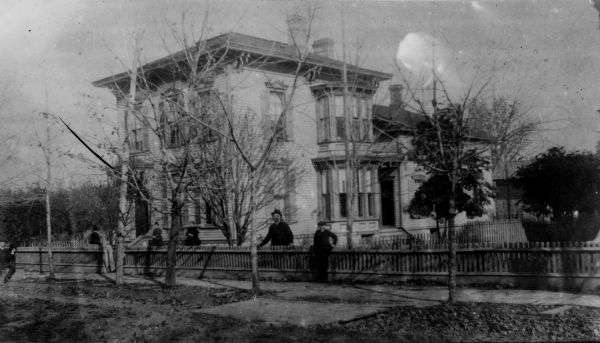 Exterior view of the residence of D.J. Morey, with multiple individuals standing in and around its front yard. A fence runs along the sidewalk.