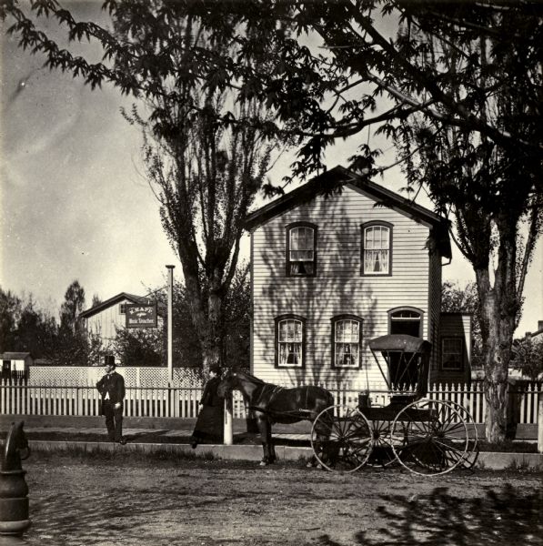 Exterior view of the home of M. Happ, with a horse-drawn wagon parked along the curb, and a man and woman standing on the terrace. A sign next to the house is on the right and reads: "M. Happ, Music Teacher." In the left foreground is a horse head hitching post.