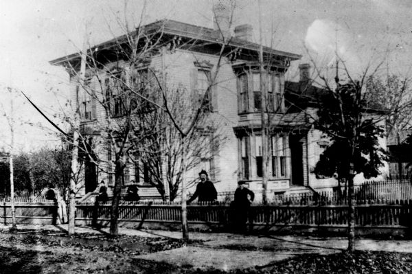 Elliott home, residence of Hames T. Elliot, at 7th Avenue. People are posing at the fence, and near the entrance.