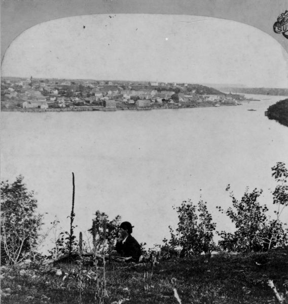 Half stereograph of an elevated view from hill showing the confluence of the Mississippi River and the St. Croix River. A man wearing a hat is in the foreground looking to the left, and he is sitting or standing just below the edge of the hill.