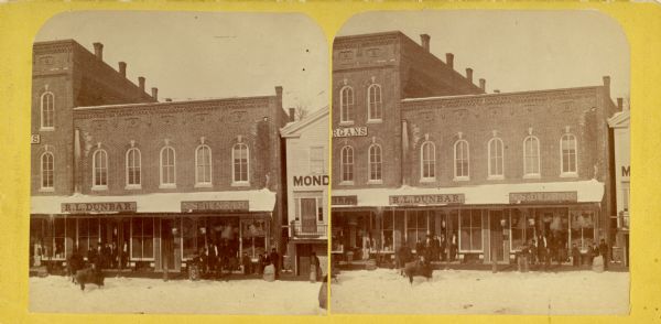 Stereograph view of several establishments along Main Street with people standing along the walkway in front of them. The signs on the buildings read: "R.L. Dunbar" and "N.S. Dunbar."