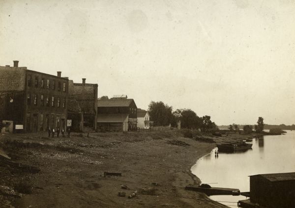 View along a levee, with buildings on the left. Dunbar's Hall, the large building in the foreground, burned down during the winter of 1907-1908. Five men are standing on the left in front of one of the buildings. There are signs for the Minnesota State Fair on a building behind them. On the right two children are standing in shallow water near a barge.