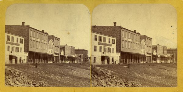 Stereograph view of the levee, with buildings along the left. Signs on the buildings read, left to right: "(?) Hotel", "Saloon" and "Dunbar's Hall." People are standing in front of the buildings.
