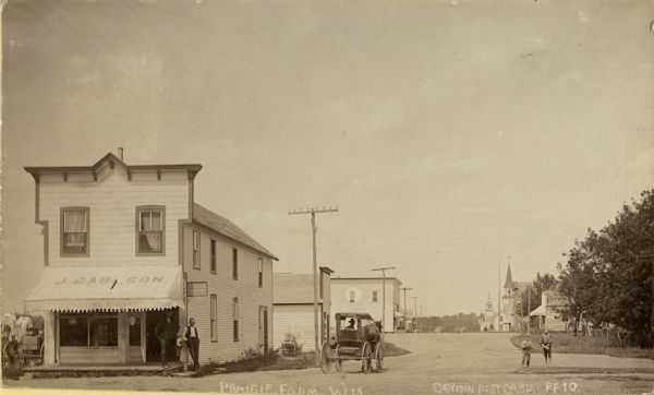 View of several buildings in Prairie Farm. Men are standing under the awning of the J. Carlson storefront on the left, a man is driving a horse-drawn wagon in the road, and two boys are standing in the road on the right. Commercial buildings are further down the road.