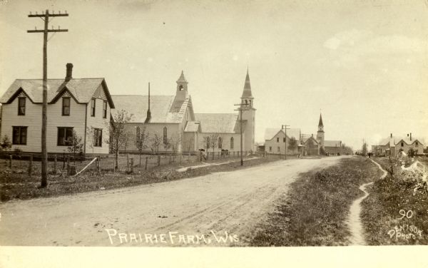 View of an unpaved street in Prairie Farm, lined on the left with multiple buildings, including three churches. Along the right side of the road is a dirt path.