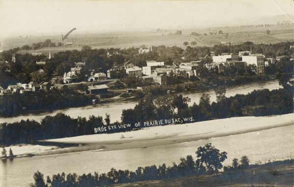 Elevated view of buildings lining the river in foreground, with farmland in background. Caption reads: "Bird's Eye View of Prairie Du Sac, Wis."