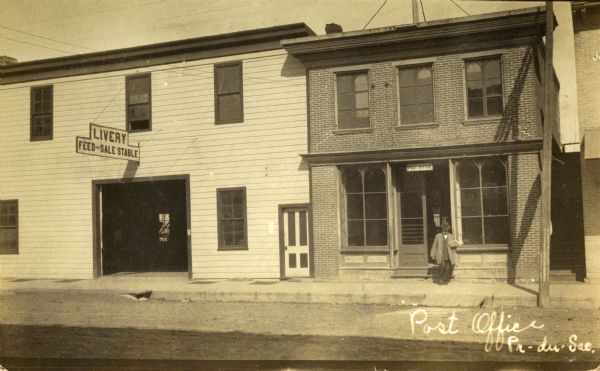 Two buildings: a post ofice and a livery stable/feed store. A man is exiting the post office. Sign on livery stable reads: "Livery Feed and Sale Stable." Caption reads: "Post Office Pr-du Sac."