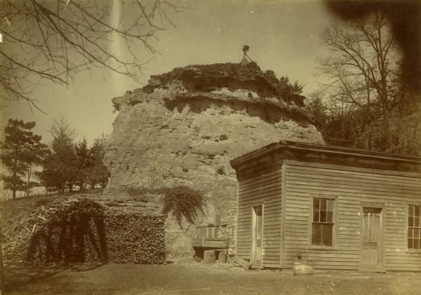 Old Uncle Billy Johnson home at Governor Phillip's house, about six miles north of Prairie du Sac near highway 12. Portion of house, rock formation in background, and large stack of firewood.