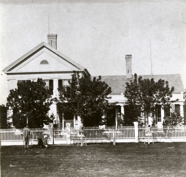 View towards the front of a house with a white fence. Posing in front of the fence are a man (appears to be on crutches), a young child, and a dog. Other people are posing behind the fence and on the porch.