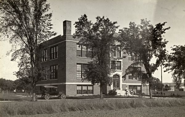 View from field towards the high school. There is a small group of people sitting on front steps, and an automobile is parked in front on the left. Houses are in the background on the right.
