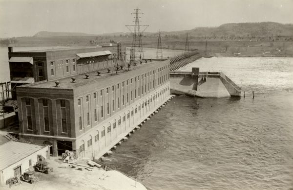 Elevated view of dam and power station on a river. Two automobiles are parked in a lot in the lower left corner. Bluffs are in the distance.