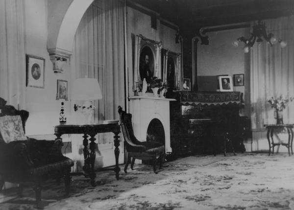 Image of the drawing room on the east side in the Spaulding house. The room was furnished by Marshall Field and Co. in 1870.<p>East end of the room, showing the piano, which is now in the Methodist Church hall.  The original wallpaper is still on the room and the original carpeting, pink roses on a grey background. The furniture is of walnut. Over the mantle on the marble fireplace is an oil portrait of the late D.J. Spaulding, and at its side a smaller picture of his nephew, the late May Price, who was a favorite of the family.  The west end of the room has an oil painting of the Mrs. Spaulding on the wall.<p>Misses Mary and Hane Spaulding sold the home in 1949 to E.E. Homstad and Ralph Boehlke. The two sisters moved out in 1943 to an apartment and the house was not lived in for some years. The furnishings are still owned by the Misses Spaulding and Mrs. Sidney Castle, a sister.