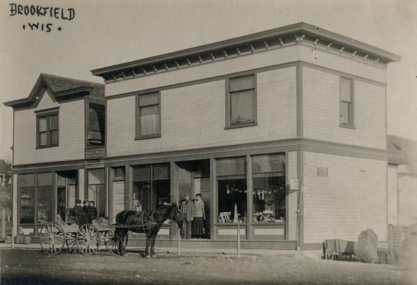 View of storefront and people with a horse-drawn carriage.
