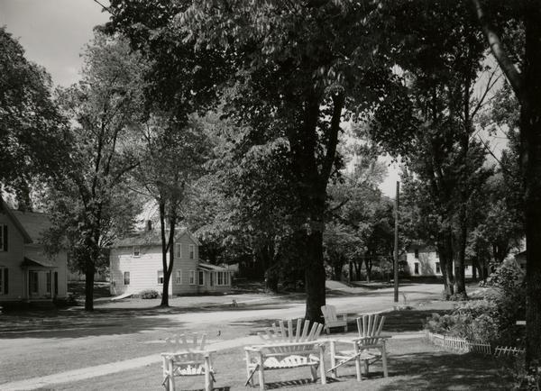A set of wooden lawn chairs are in the foreground on a lawn. They are facing a tree-lined residential street.