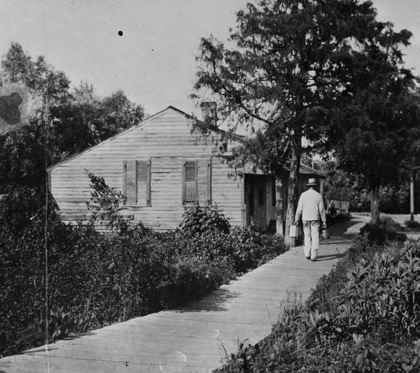Side view of the John Davis residence, the first frame house in Burlington, with plank sidewalk. A man is walking away down the sidewalk.