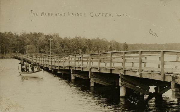 View of the Narrows Bridge, with a group of people on the left sitting and standing in a boat. The far shoreline is tree covered. Caption reads: "The Narrows Bridge Chetek, Wis."