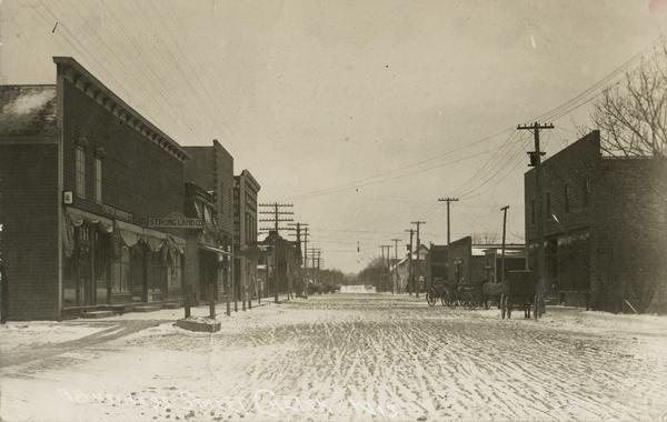 View down a snow-covered unpaved street in Chetek. Horse-drawn vehicles are along the right, and commercial buildings line both sides of the street. There is a sign for the "Strong Land Co." in front of a storefront on the left.