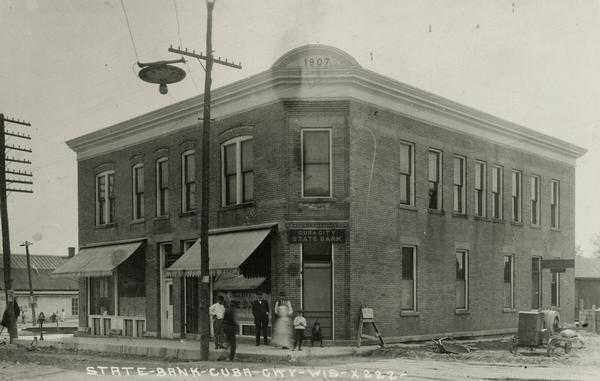 View from intersection towards the Cuba City State Bank. Caption reads: "State-Bank-CubaCity-Wis". People are standing on the sidewalk in front of the bank.