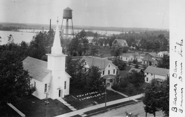 Elevated view of Cumberland with Beaver Dam Lake in the background. In the foreground is a street, and across the street is a church, a water tower and dwellings among trees. There is a man on a ladder leaning against the side of the church. Caption reads: "View of Lake, Cumberland, Wis."