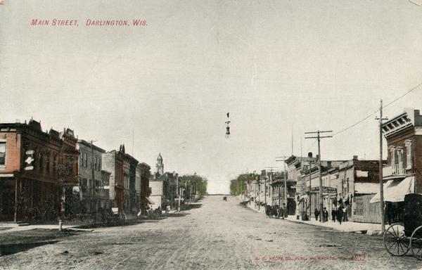 View down center of unpaved Main Street with storefronts on both sides. A group of people are on the sidewalk on the right. Caption reads: "Main Street, Darlington, Wis."