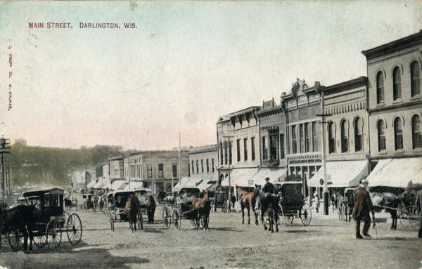 View across unpaved street towards storefronts on the right. There are horse-drawn carriages and pedestrians moving up and down the street. Caption reads: "Main Street, Darlington, Wis."
