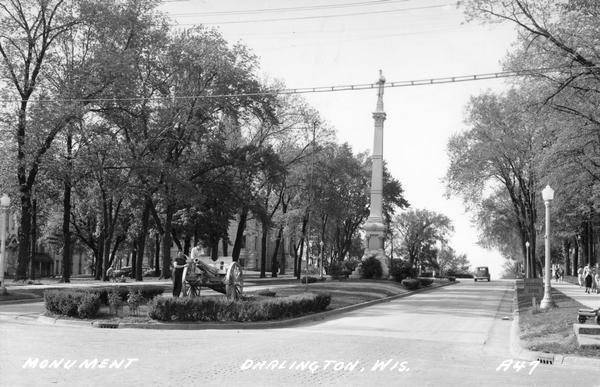 View looking up street towards the Soldiers' Monument on the median. People are standing near a cannon in the median in the foreground. Caption reads: "Soldiers' Monument, Darlington, Wis."