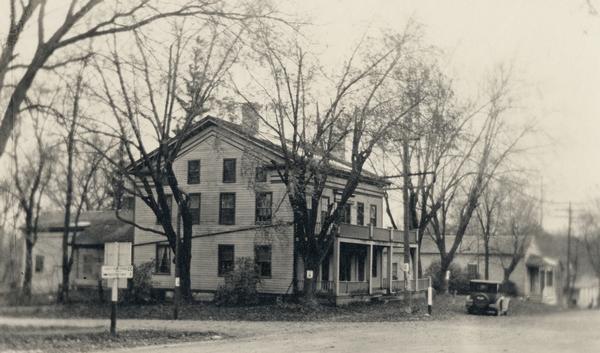 A car is parked in front of Hawks Tavern, also known as Hawks Inn, with trees, a street sign and power lines.