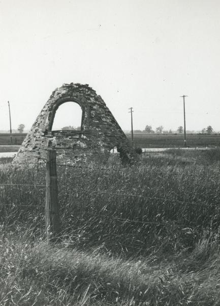 Charcoal kiln in ruins in a field beyond a fence, with electric power lines in the background.