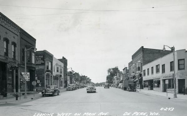Caption reads: "Looking West on Main Ave." View down center of commercial area, with automobiles moving down the street, and parked along the curbs. A U.S. flag is flying from a flagpole on the sidewalk on the left near a storefront with a sign that reads: "Milwaukee Gettelman Beer Dukes". On the right is a U.S. flag on the sidewalk in front of the Post Office. A water tower is in the background on the right just above trees.