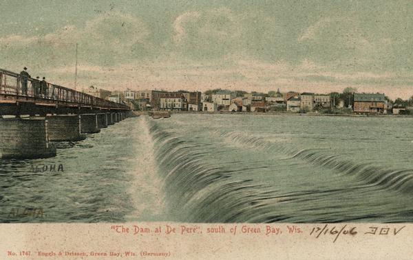 The caption reads: "'The Dam at De Pere', south of Green Bay, Wis." View of the dam towards the far shoreline, with pedestrians walking on the bridge above on the left.