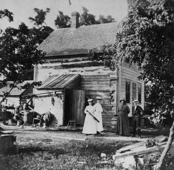 A group of people, men and women, standing outside in the yard in front of Eleazer William's log cabin, five miles south of De Pere. The women in the center is holding a long object, perhaps a firearm.